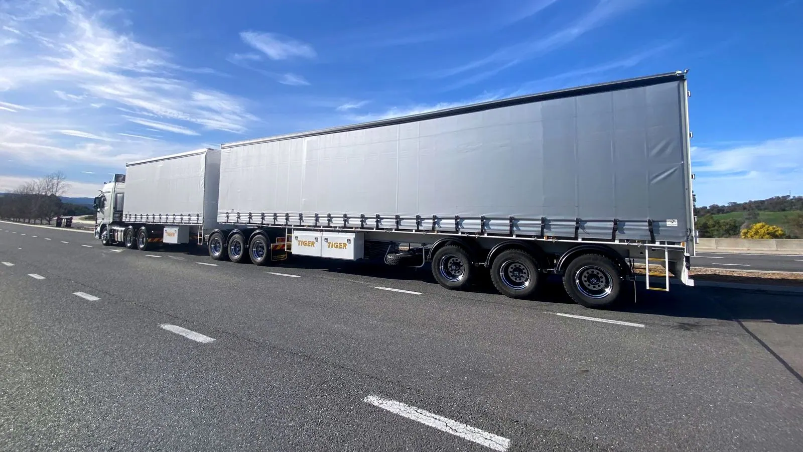 A Digiton B-Double seen side on, both curtain-side trailers coupled, parked on the shoulder of a highway.